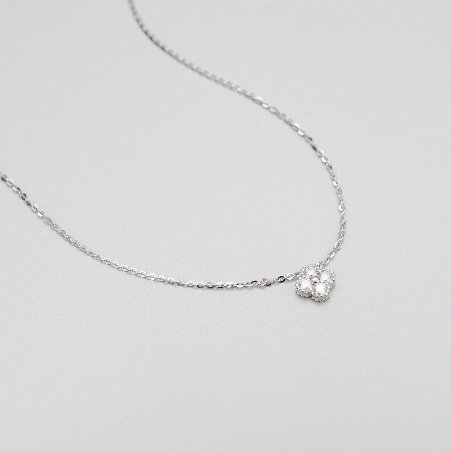 A delicate silver chain necklace featuring a small, textured silver heart-shaped pendant with a clear central gemstone, displayed on a neutral light gray background.
