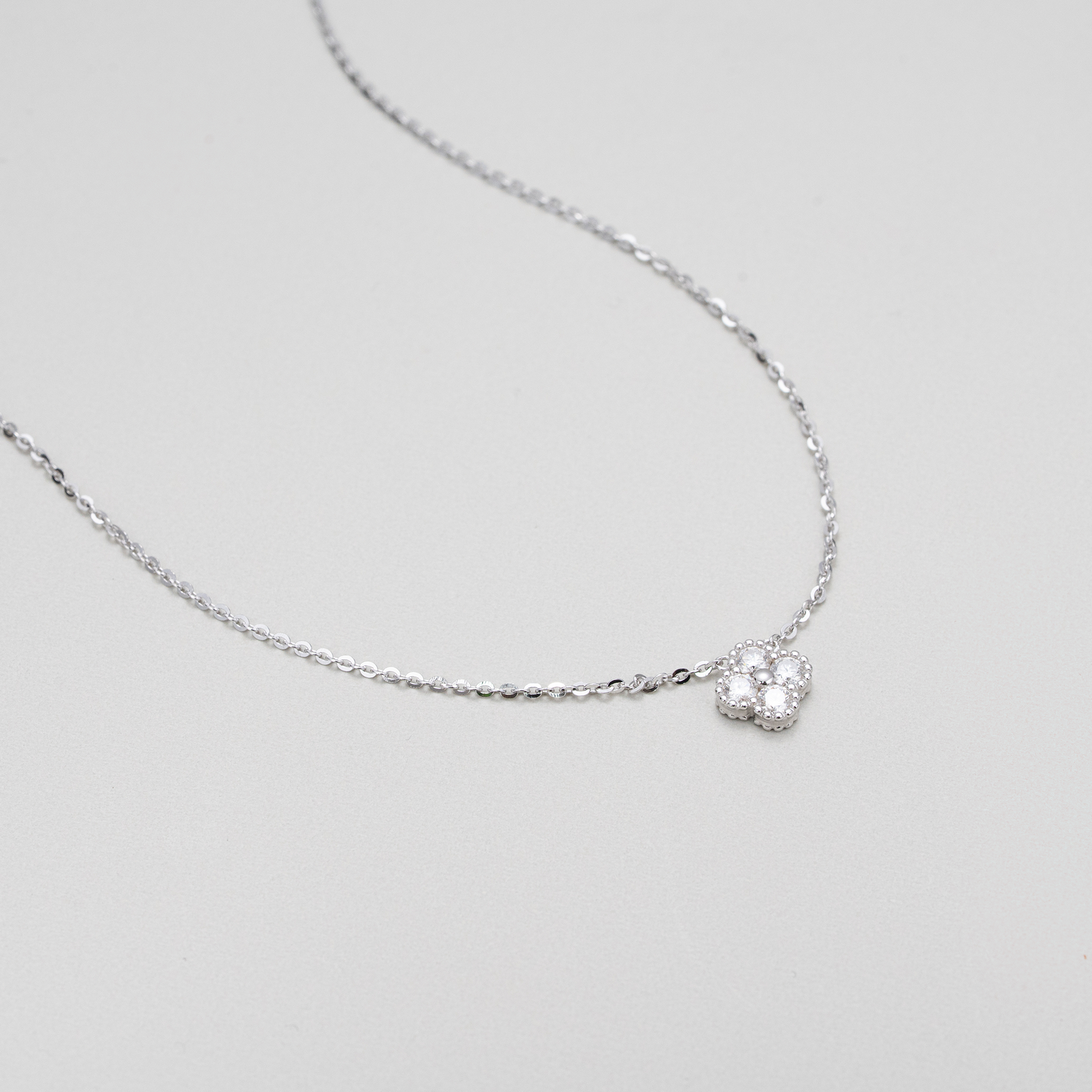 A delicate silver chain necklace featuring a small, textured silver heart-shaped pendant with a clear central gemstone, displayed on a neutral light gray background.