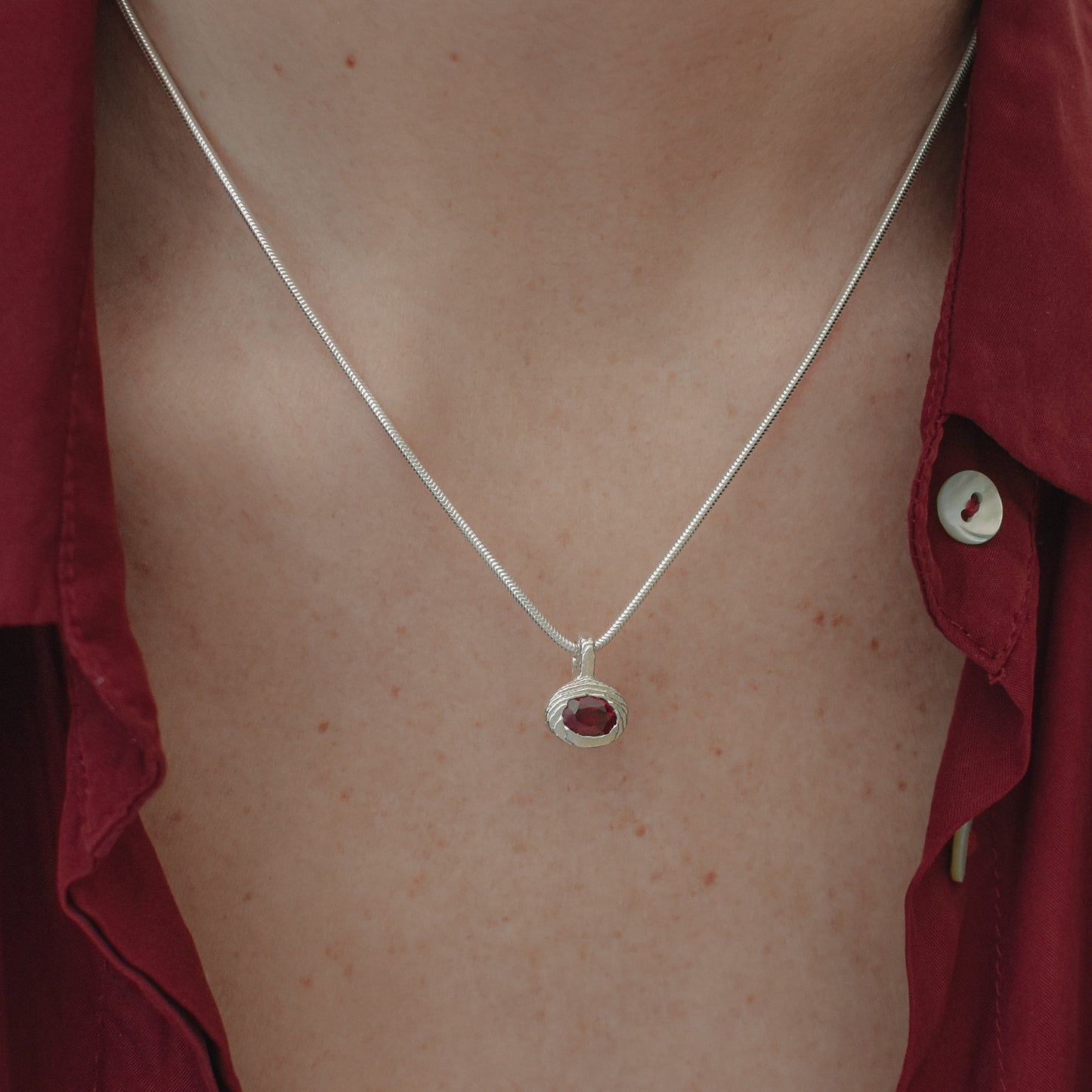A close-up of a person's chest wearing a delicate silver chain necklace with a small oval red gemstone pendant, styled with an open dark red button-down shirt.