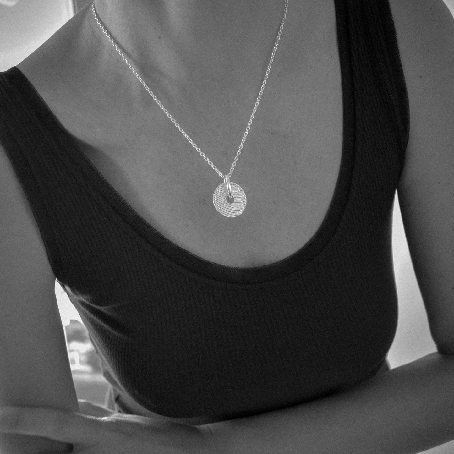A close-up of a person wearing a delicate silver chain necklace with a textured circular disc pendant, paired with a black ribbed tank top.