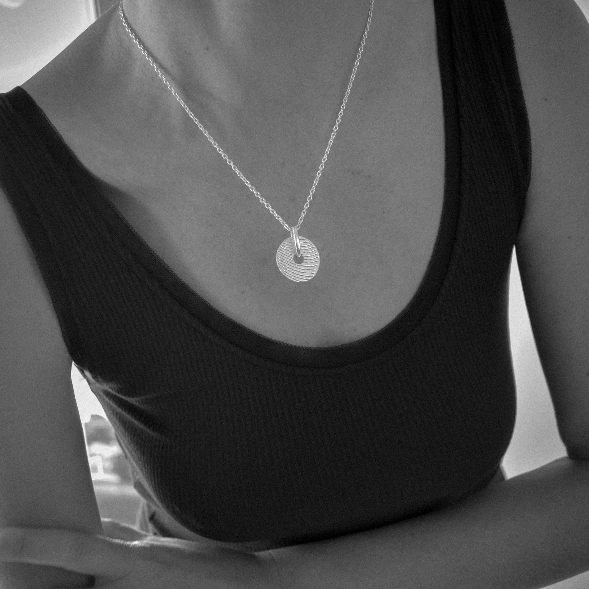 A close-up of a person wearing a delicate silver chain necklace with a textured circular disc pendant, paired with a black ribbed tank top.