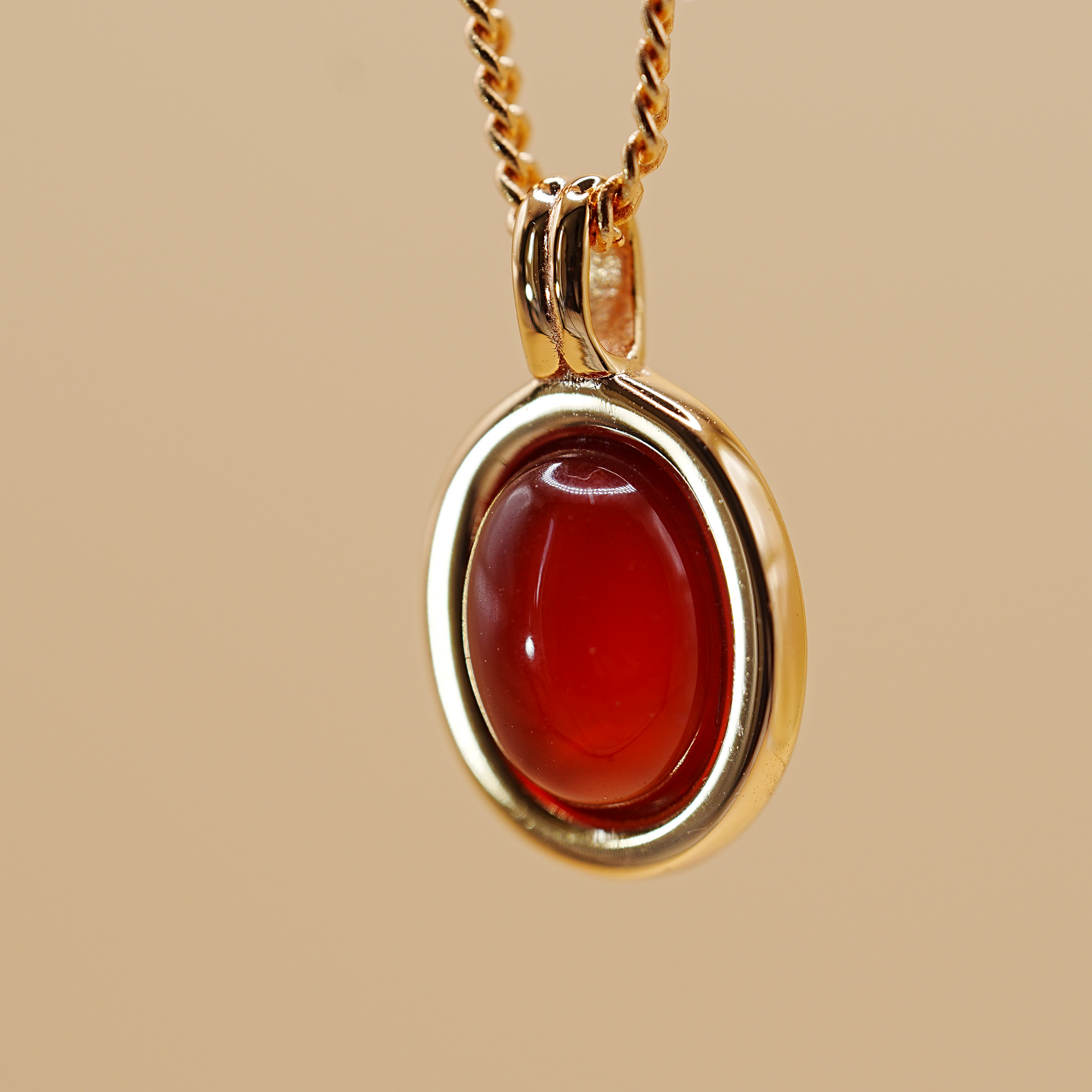A gold pendant necklace featuring a polished oval red gemstone, suspended from a gold chain against a solid beige background.