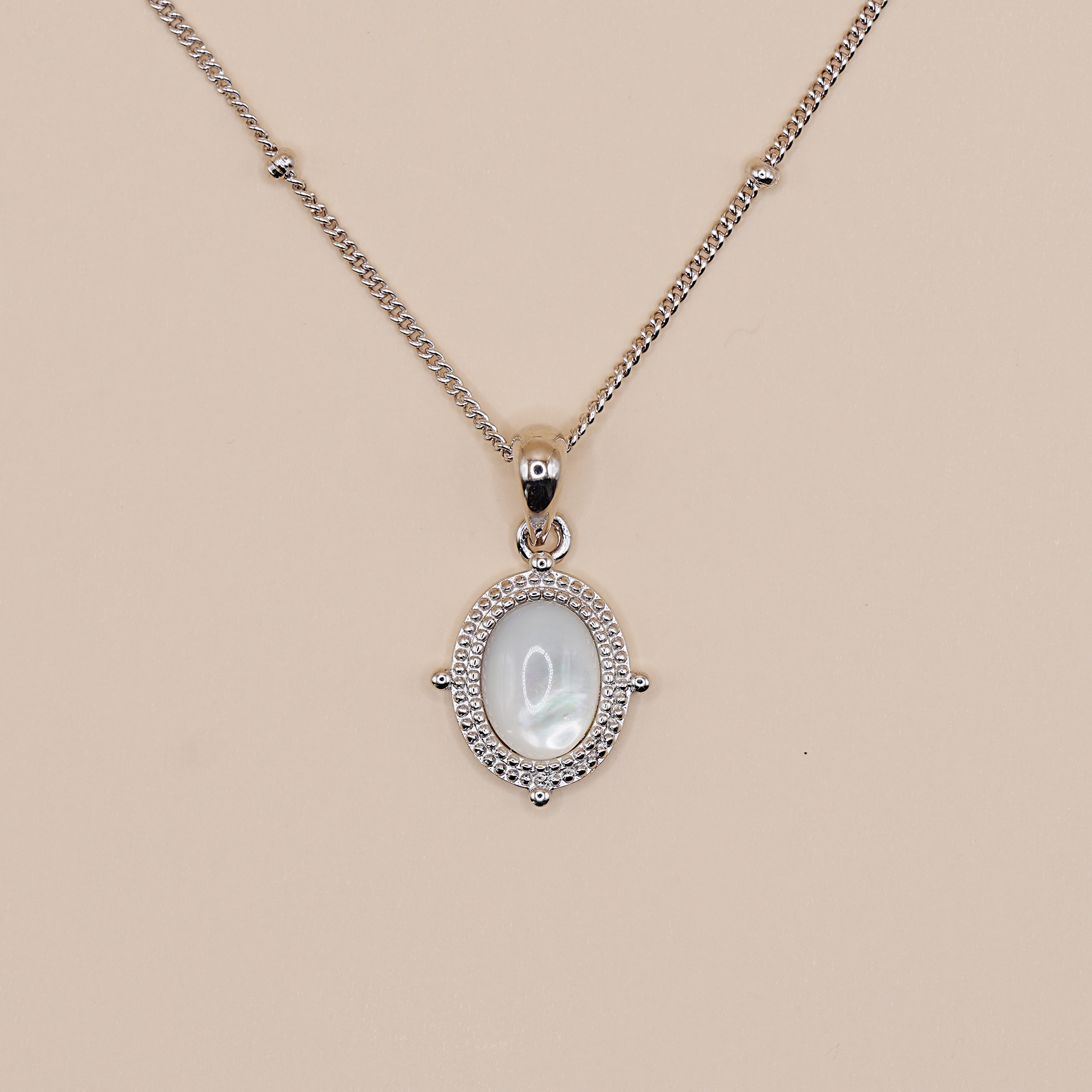 A silver chain necklace with an oval pendant featuring a white mother-of-pearl center stone surrounded by a halo of small clear stones, displayed on a solid beige background.