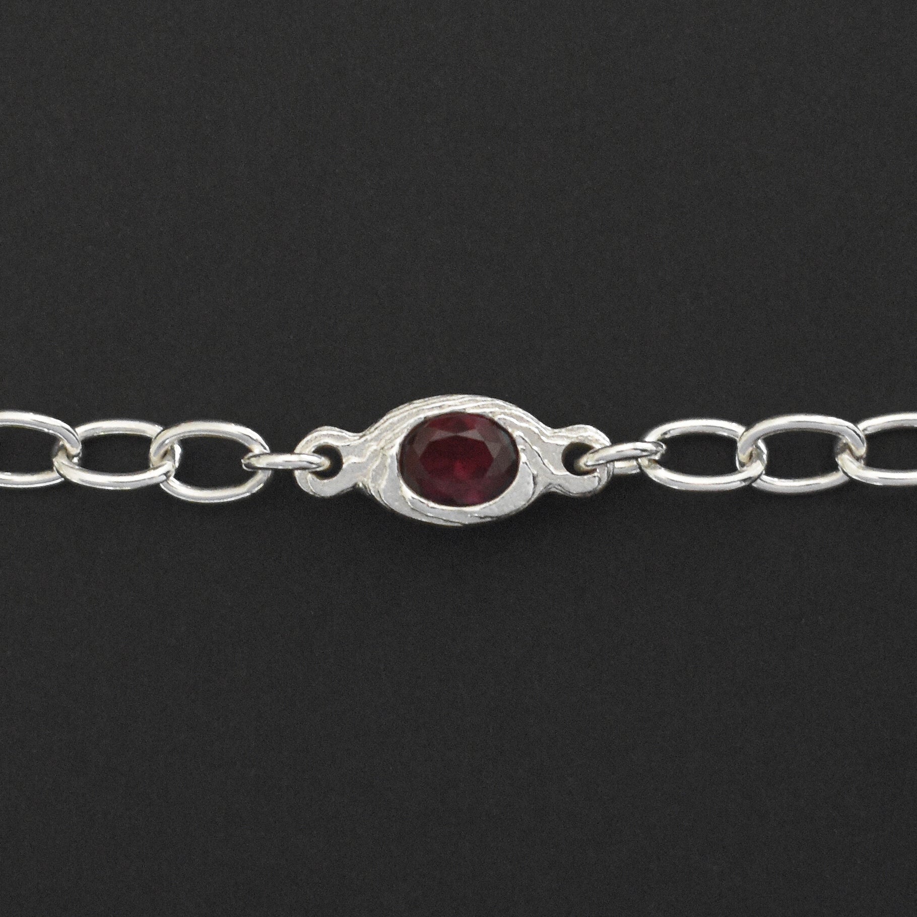 A sterling silver chain bracelet featuring a central oval bezel-set dark red gemstone, shown against a solid black background.