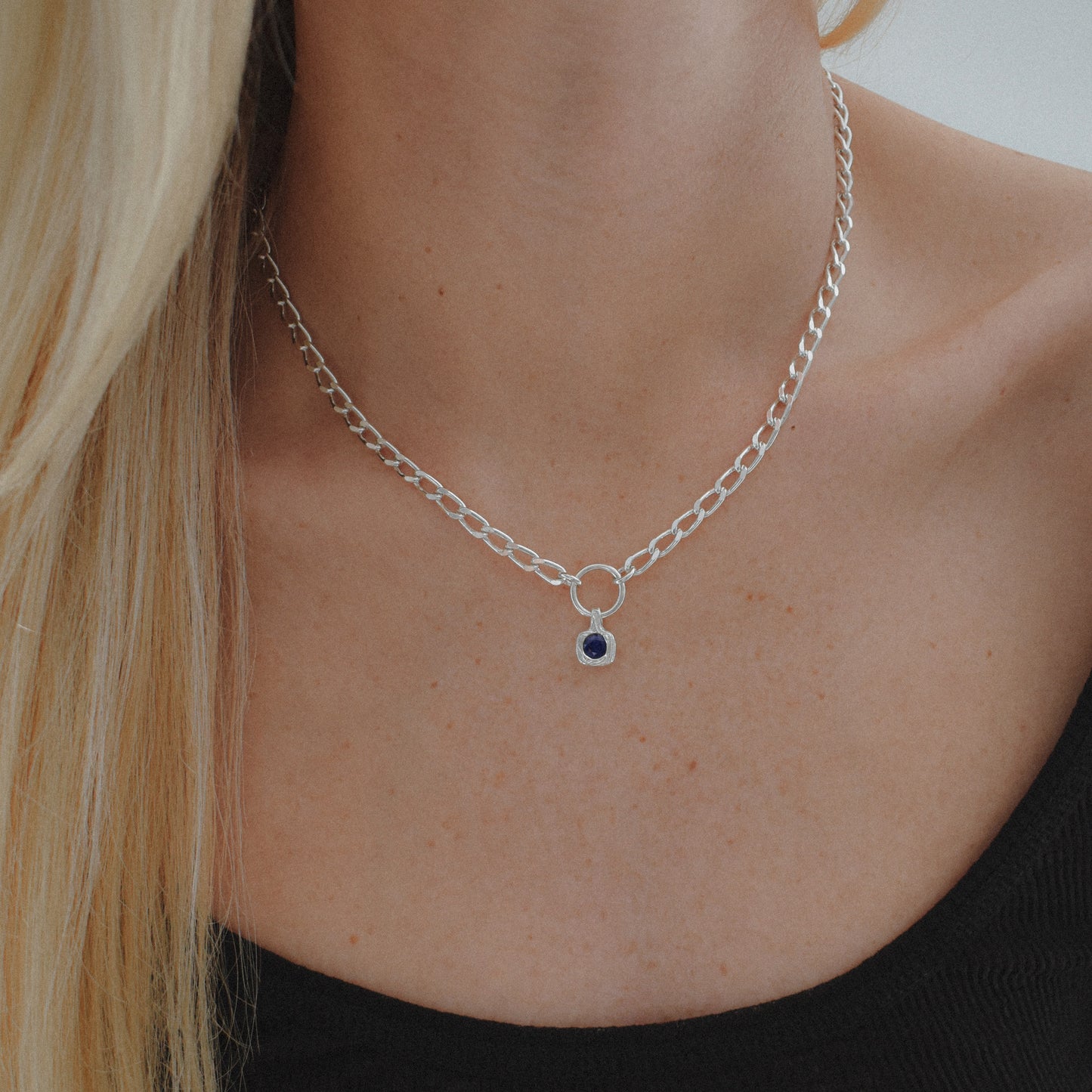 A close-up of a model's neck and chest wearing a silver chain necklace with a small circular ring centerpiece and a tiny dark blue oval gemstone.