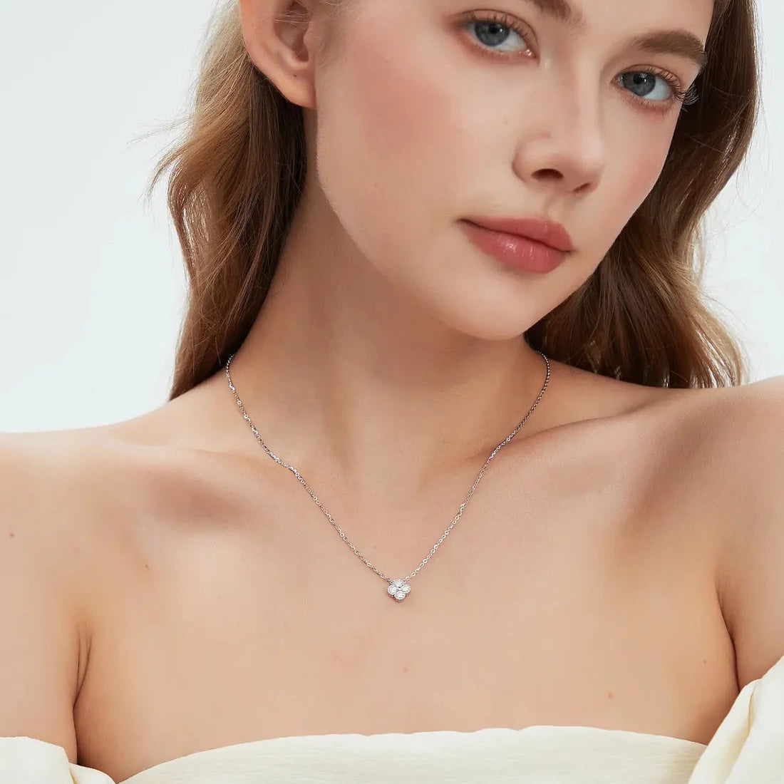 A close-up of a woman wearing a delicate silver chain necklace with a small, clear, heart-shaped gemstone pendant. She is wearing a cream-colored strapless top against a clean, light background.
