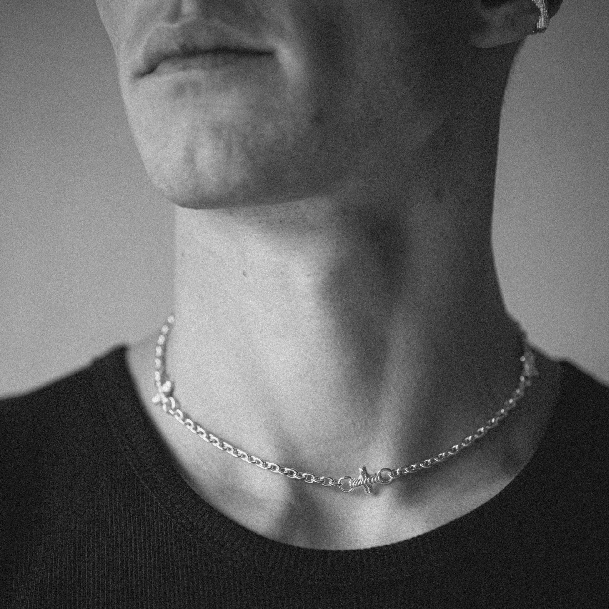 A close-up black and white photograph of a man's neck and lower face wearing a delicate silver chain necklace with a small central charm.