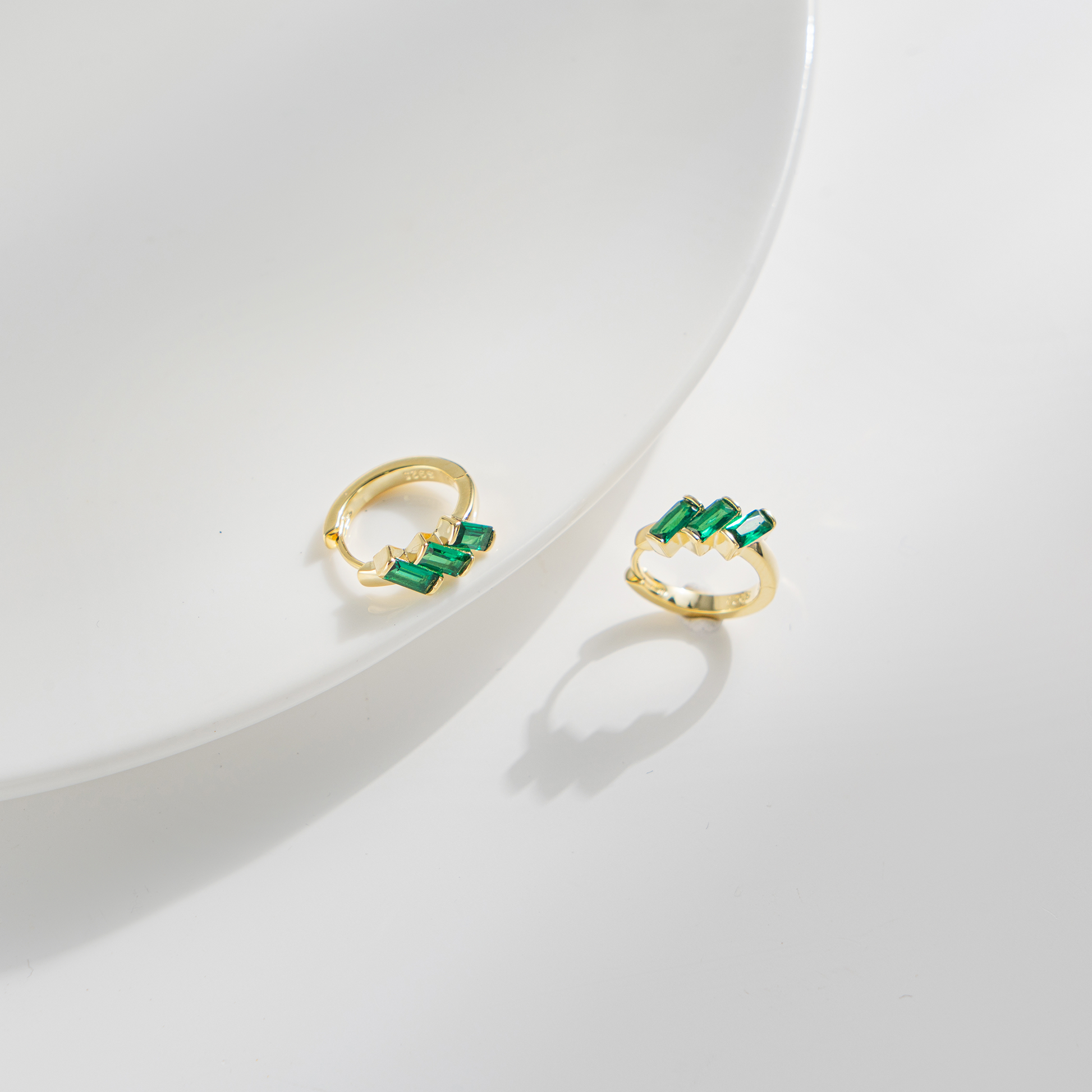 A pair of gold huggie hoop earrings with green baguette-cut stones, displayed on a clean white surface with soft shadows.