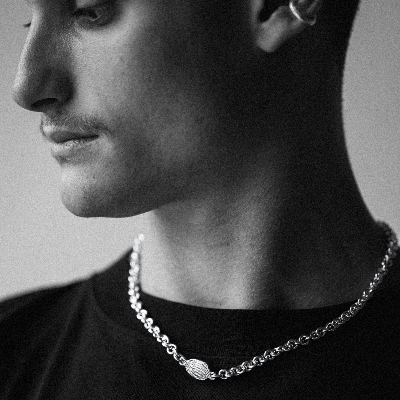 A close-up black and white photo of a man's profile wearing a silver chain necklace with a textured oval pendant.