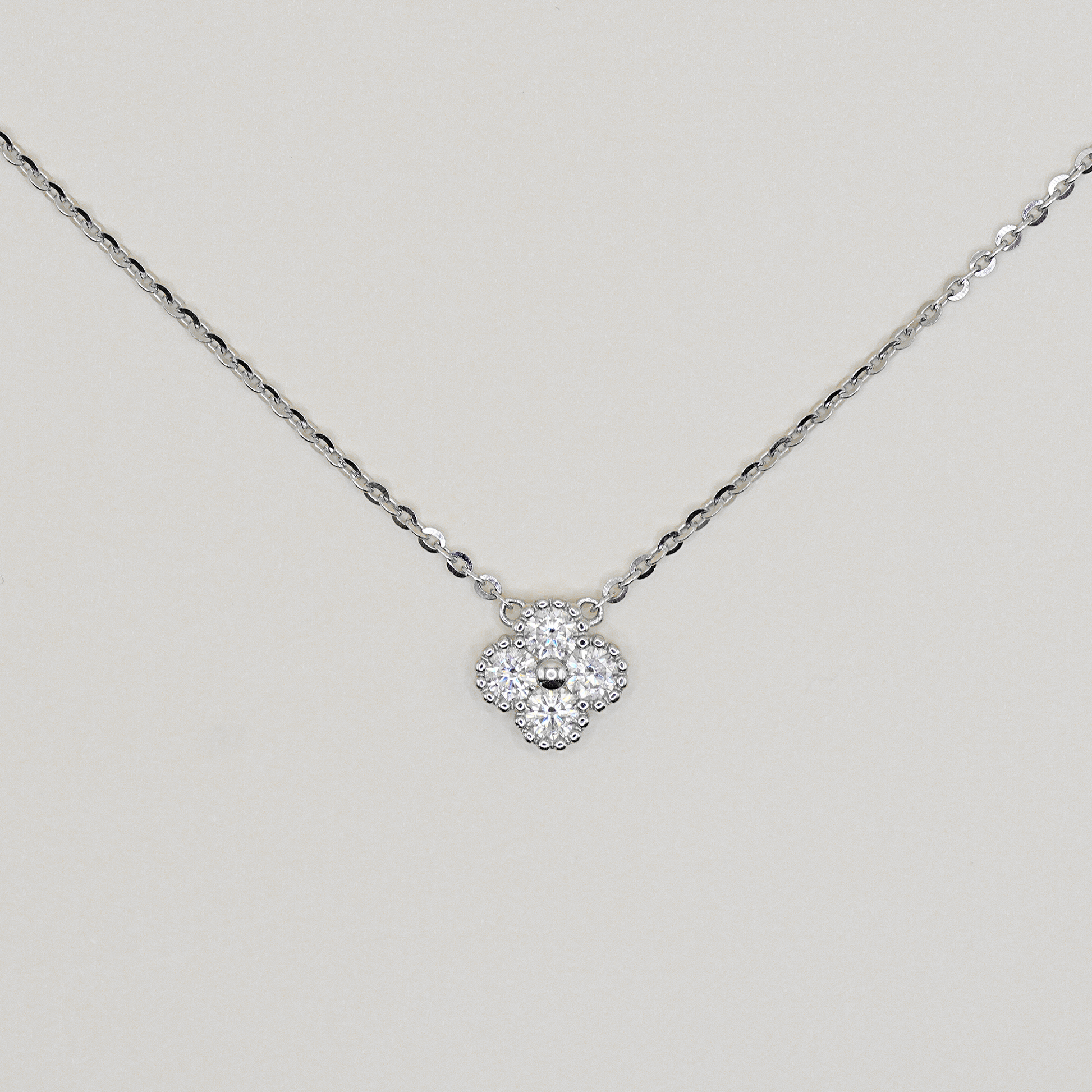A delicate silver chain necklace with a small four-petal flower pendant paved with clear stones, displayed against a light gray background.