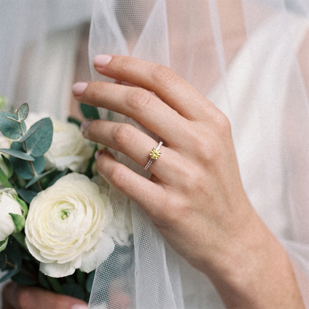 A close-up of a woman's hand wearing a delicate gold ring with a small yellow flower motif, styled with a white bridal bouquet and sheer white tulle veil.