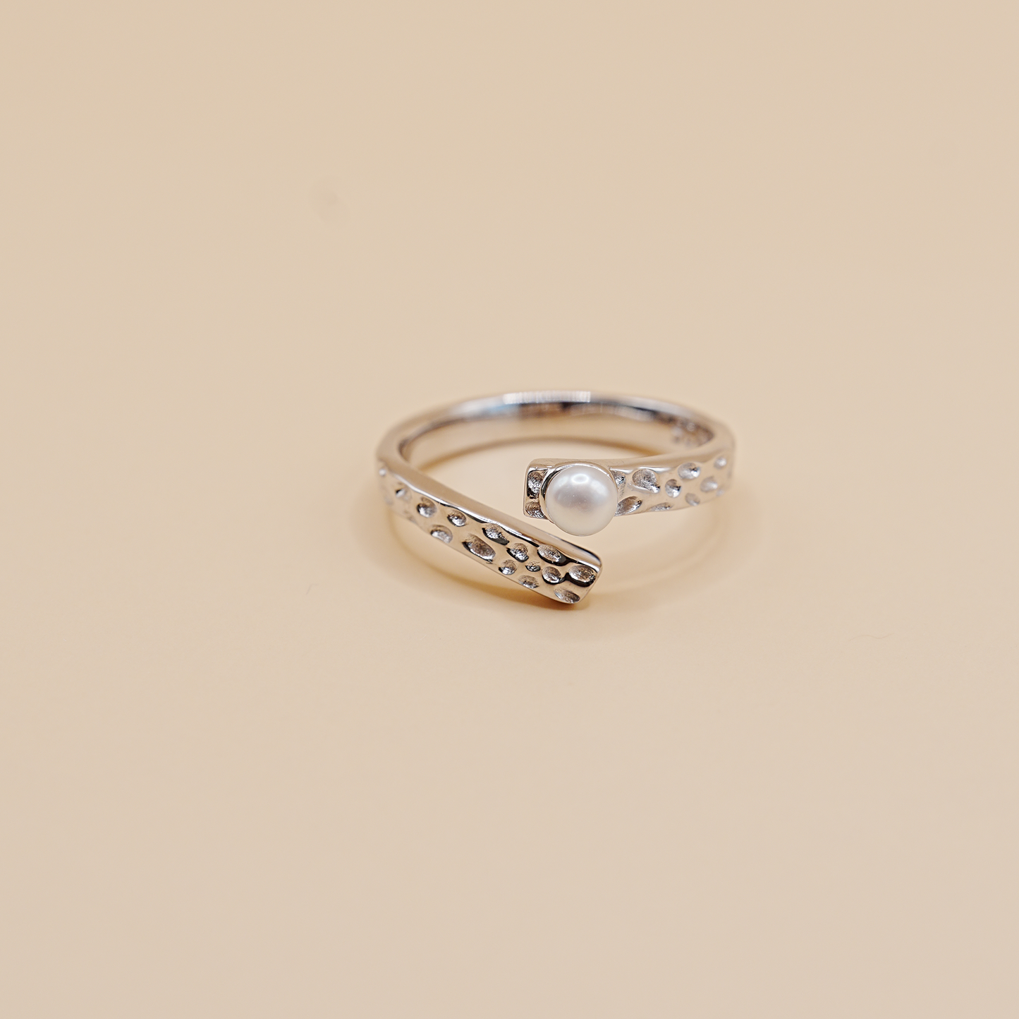 A silver open-band ring with a textured, beaded curved design and a central white pearl, set against a solid beige background.
