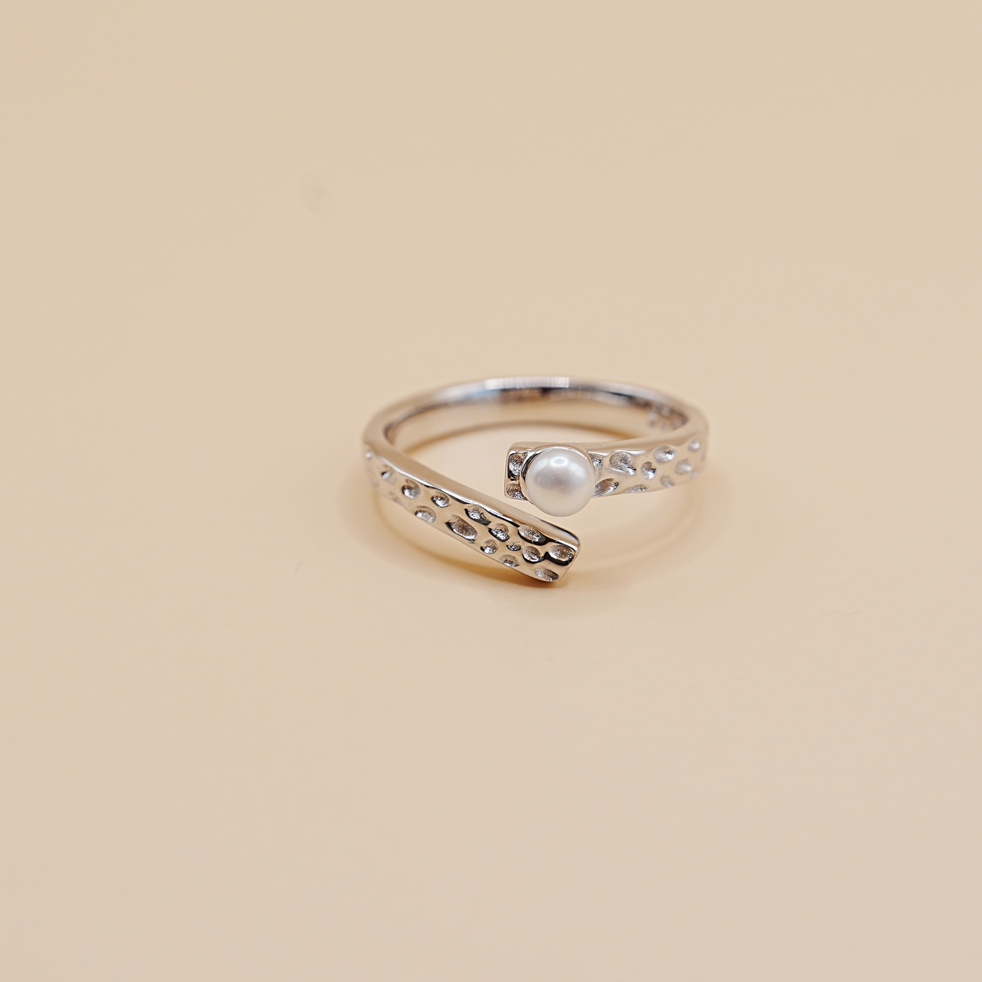 A silver open-band ring with a textured, beaded curved design and a central white pearl, set against a solid beige background.