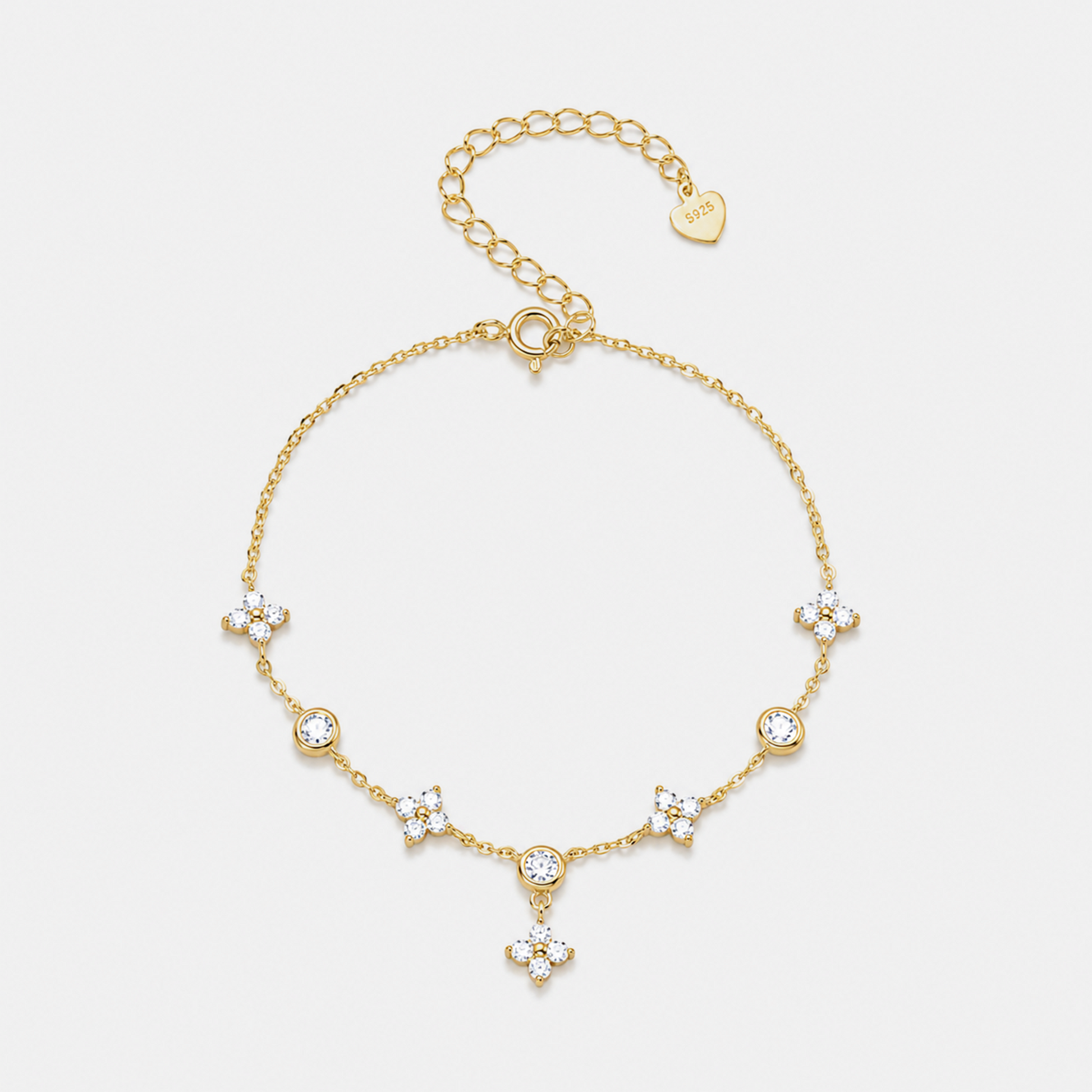 A delicate gold chain bracelet with five floral clusters of clear crystals and a small dangling crystal charm, displayed on a light gray background. A gold heart tag on the extender chain is engraved with 'SADAK'.