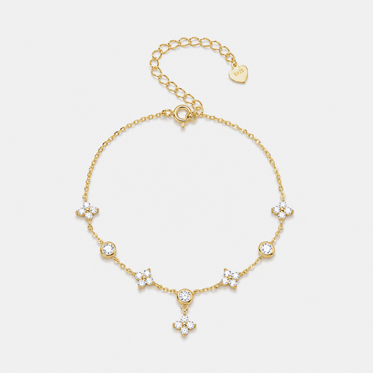A delicate gold chain bracelet with five floral clusters of clear crystals and a small dangling crystal charm, displayed on a light gray background. A gold heart tag on the extender chain is engraved with 'SADAK'.