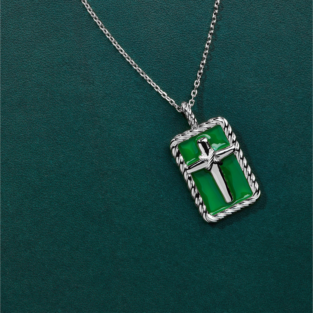 A silver-toned rectangular pendant necklace with a green enamel background and a raised silver cross in the center, hanging on a matching silver chain against a dark green textured backdrop.