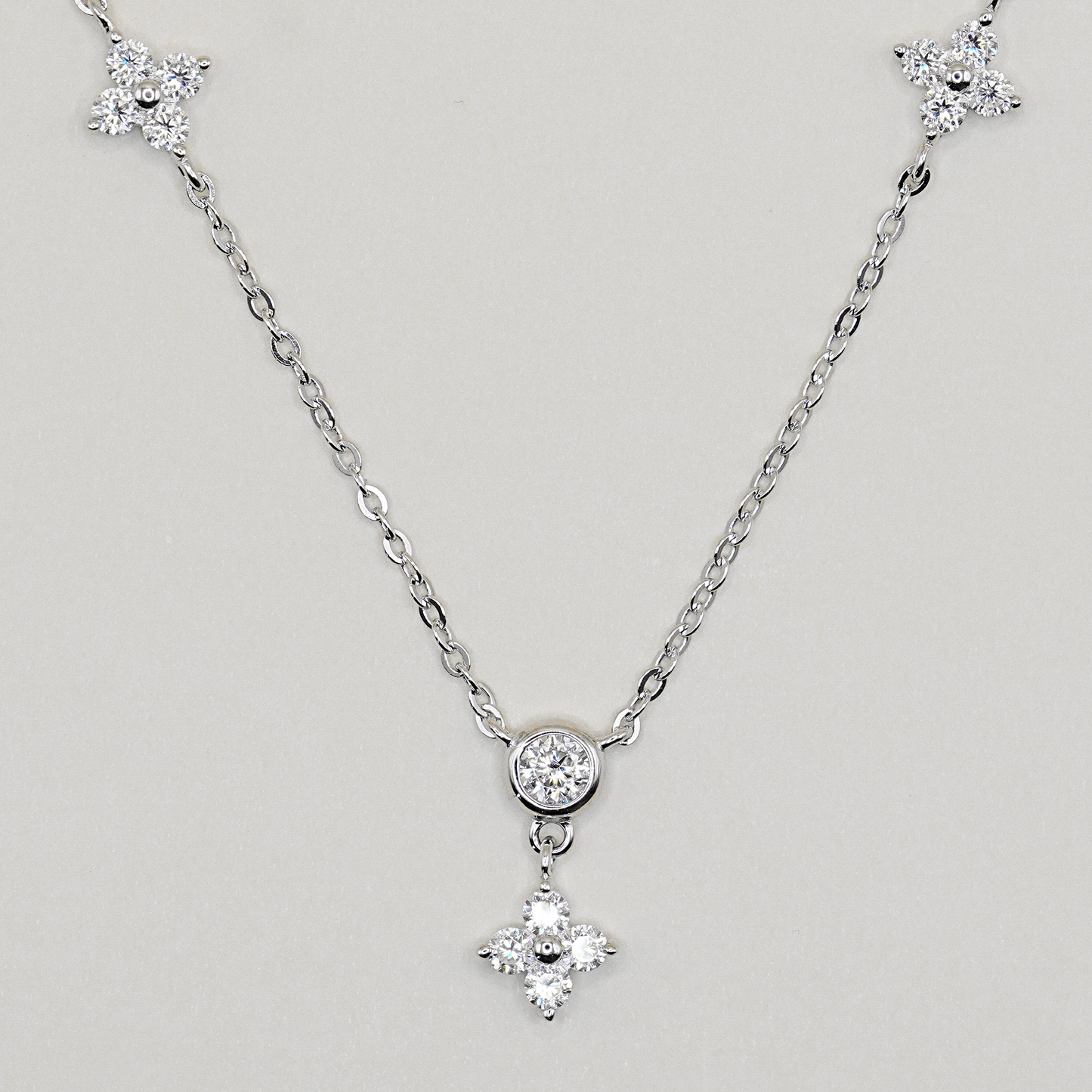 A silver-tone chain necklace featuring a central round bezel-set clear stone and a dangling four-petal flower charm, with matching flower-shaped end pieces on the chain, shown on a light gray background.