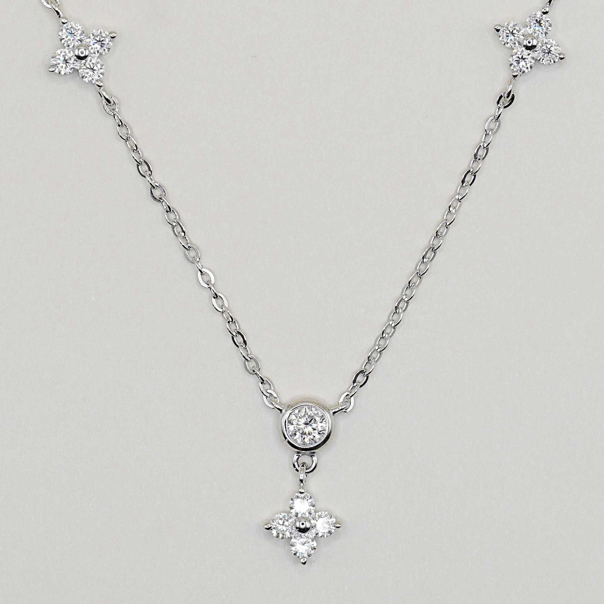 A silver-tone chain necklace featuring a central round bezel-set clear stone and a dangling four-petal flower charm, with matching flower-shaped end pieces on the chain, shown on a light gray background.