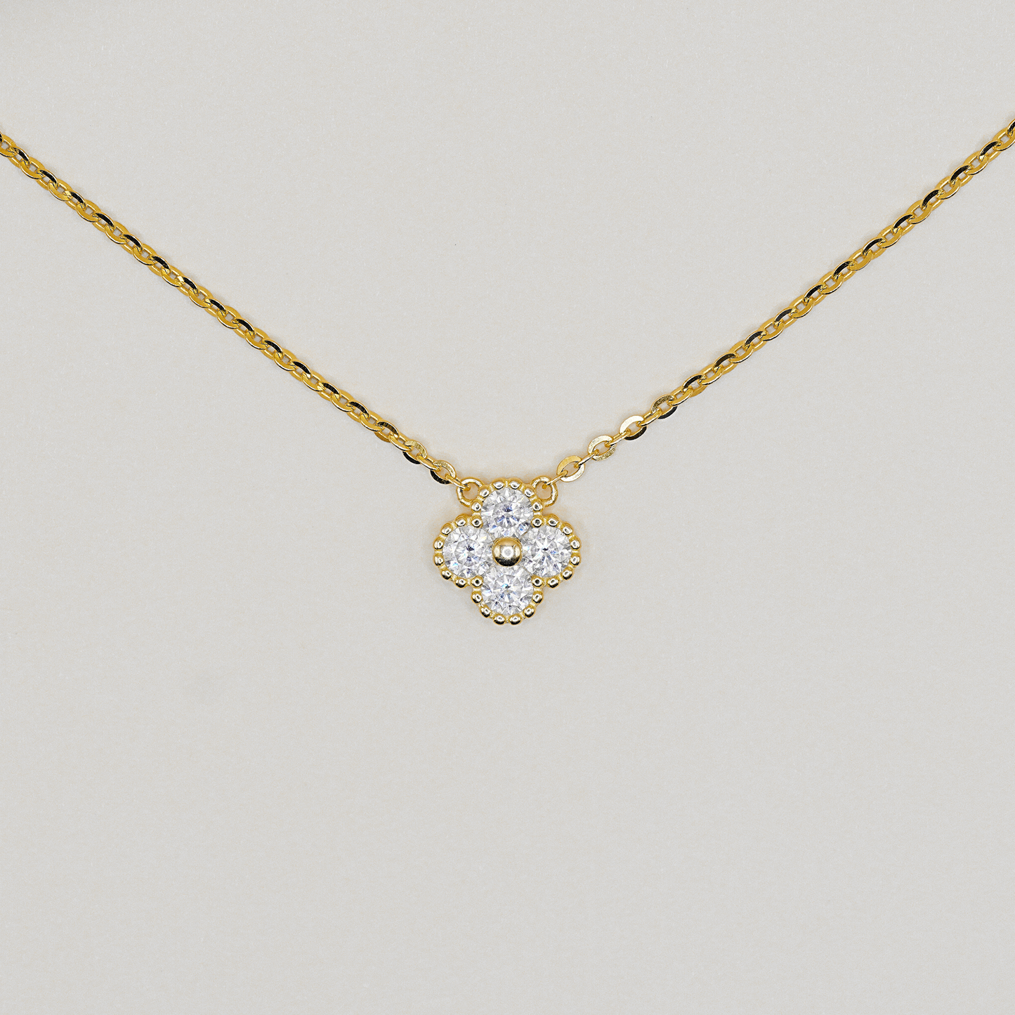 A delicate yellow gold chain necklace with a small four-petal flower pendant. The pendant is set with clear gemstones and a central gold bead, photographed against a neutral light gray background.