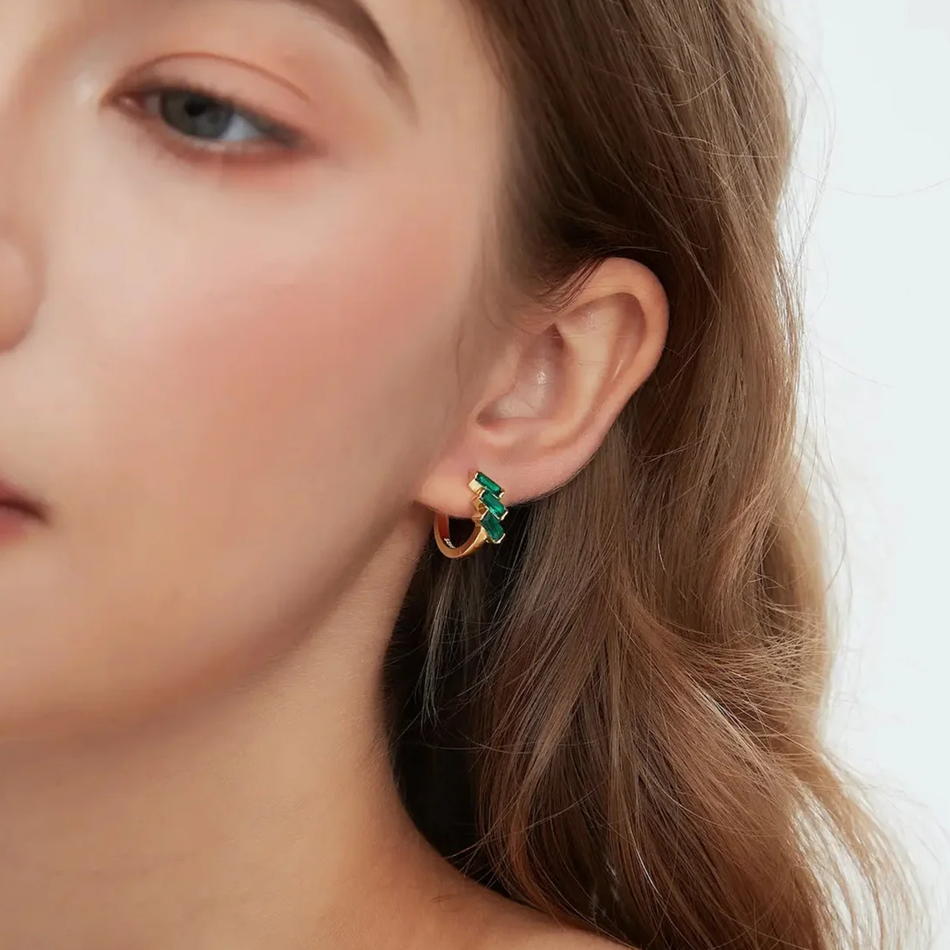 A close-up of a model's ear wearing a small gold huggie hoop earring with green enamel zigzag accents, set against a clean white background.