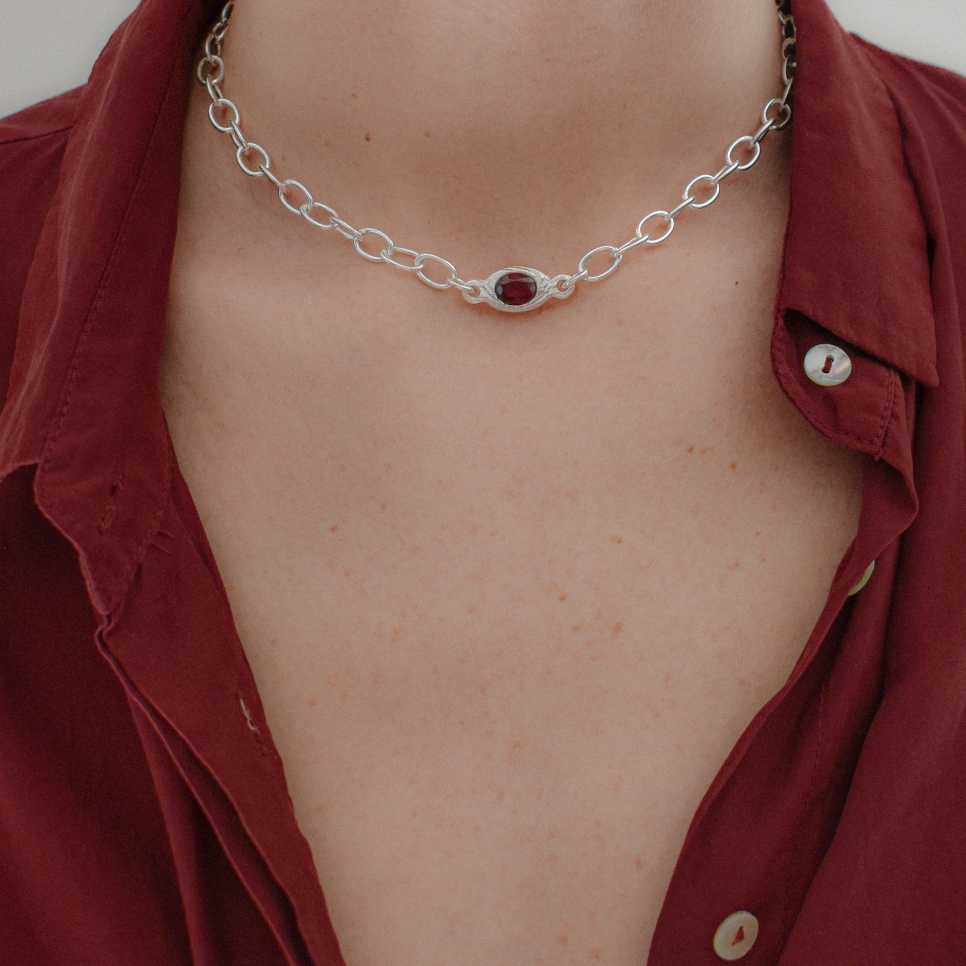 A close-up of a person's neck and chest wearing a silver chain necklace with a central oval red gemstone, paired with an open burgundy button-down shirt.