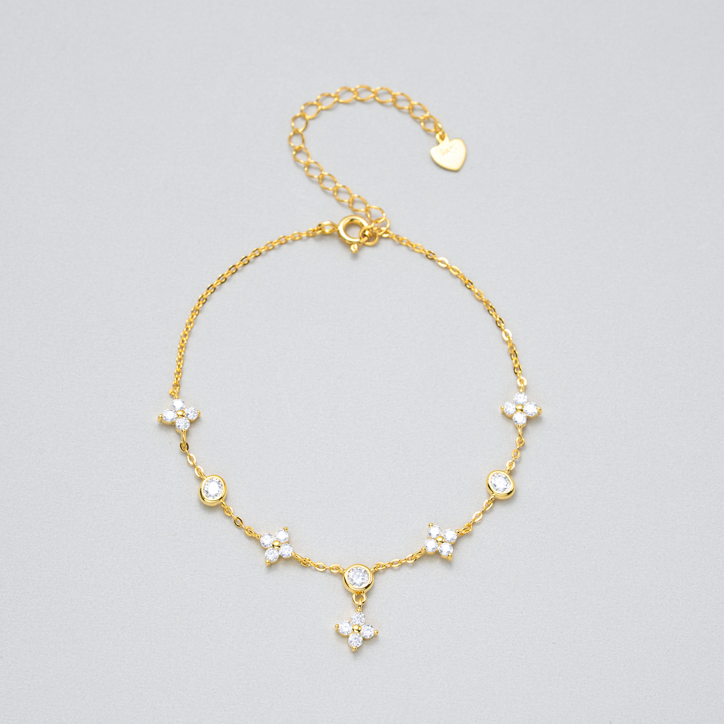 A delicate gold chain anklet featuring five small floral clusters set with clear rhinestones, a heart-shaped charm, and an adjustable clasp, displayed on a neutral light gray background.