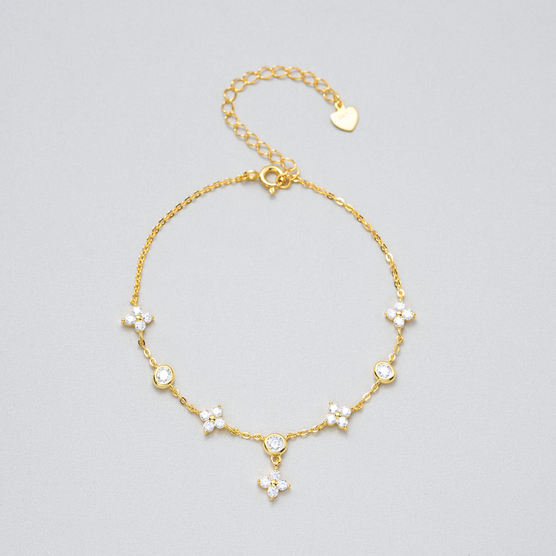 A delicate gold chain anklet featuring five small floral clusters set with clear rhinestones, a heart-shaped charm, and an adjustable clasp, displayed on a neutral light gray background.