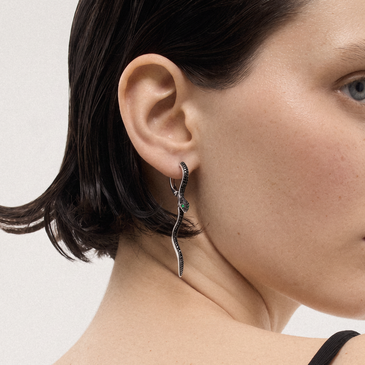 A close-up of a model's ear and neck wearing a silver wavy drop earring adorned with small black stones and a central green gemstone.