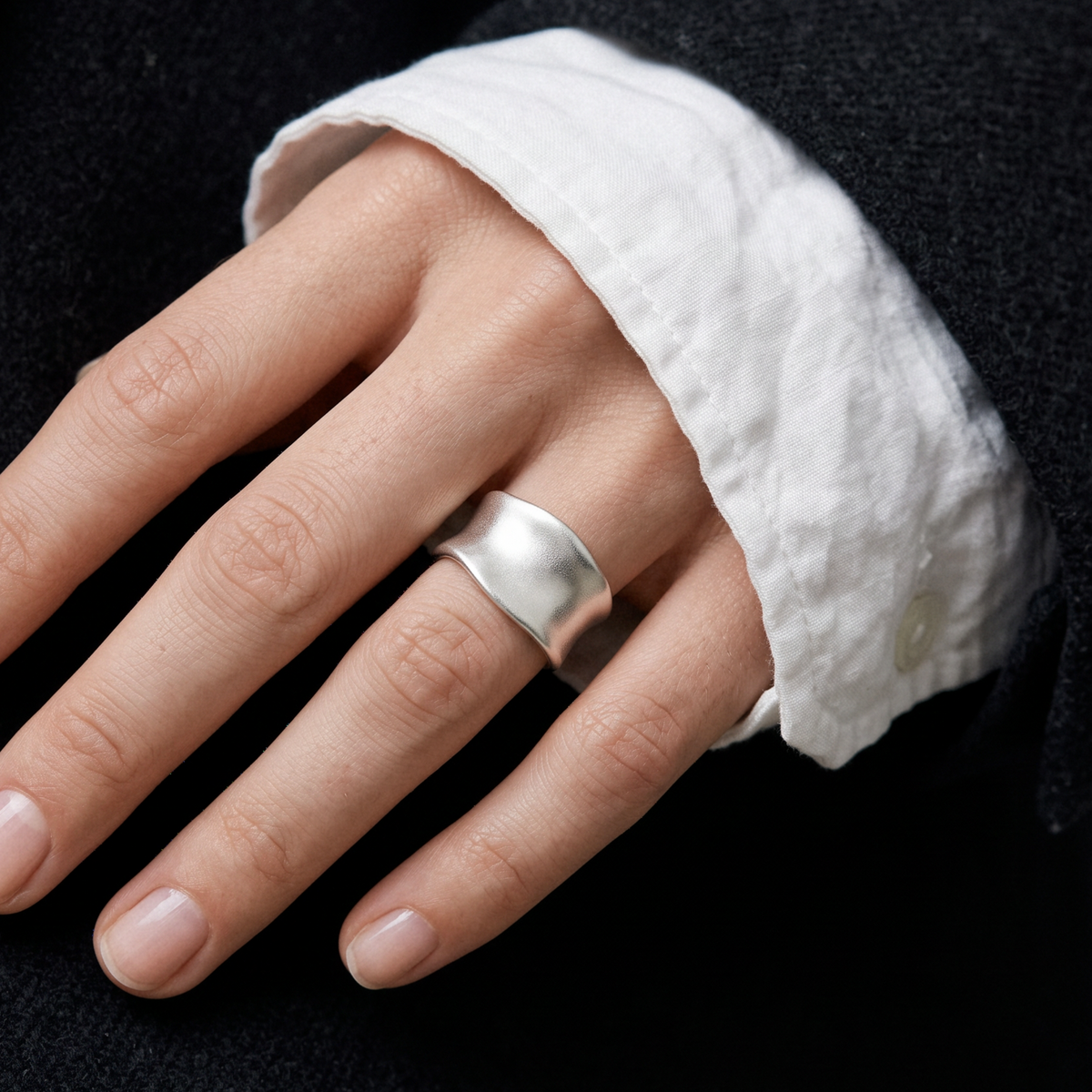 A close-up of a hand wearing a wide, polished silver band ring, styled with a white linen shirt cuff and dark charcoal jacket.
