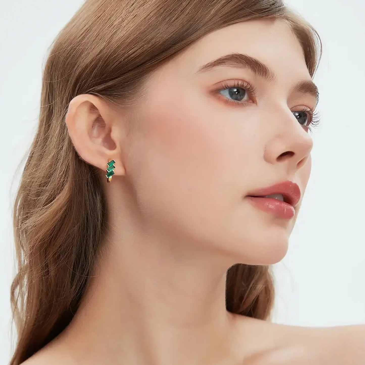 A close-up of a woman wearing a small green leaf-shaped stud earring on her ear, shown against a clean light gray background.