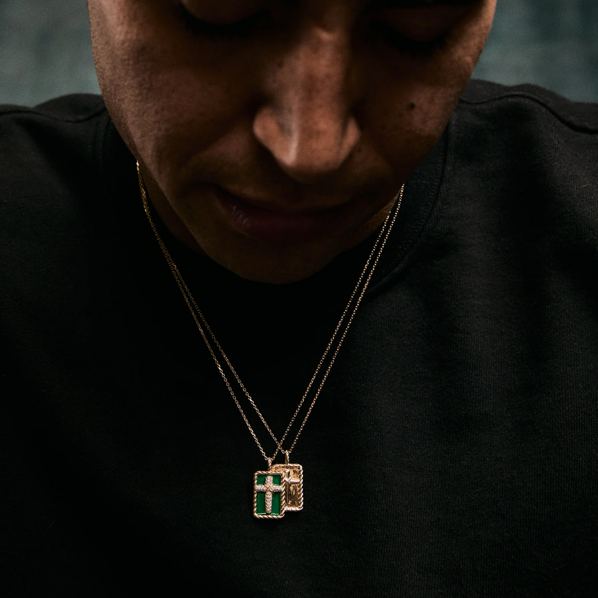 A close-up lifestyle shot of a person wearing a layered necklace with two rose gold chains and pendants, one featuring a green cross and the other a small gold cross, set against a dark black shirt.
