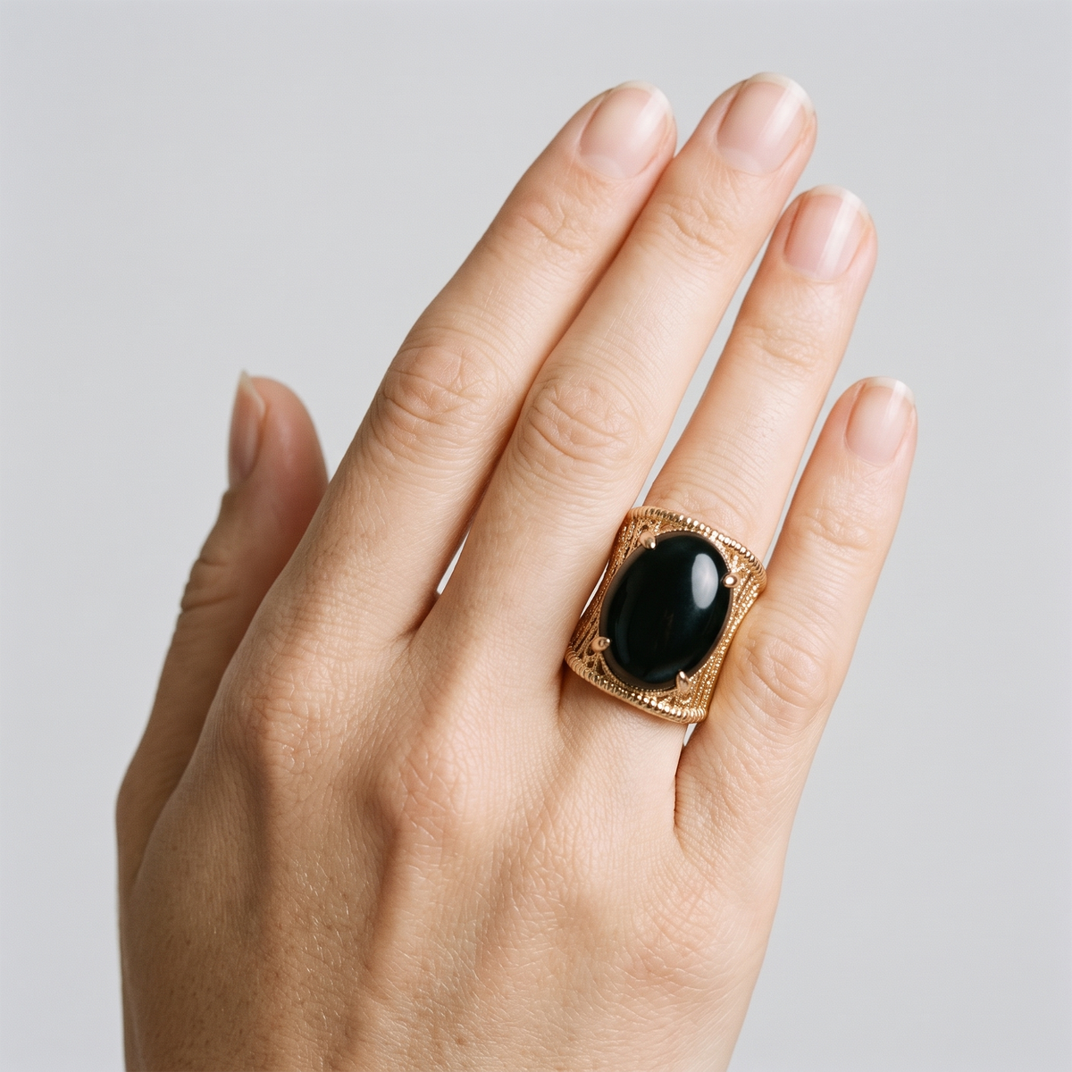 A close-up of a hand wearing a gold signet ring with a large, polished black oval cabochon stone, set against a solid light gray background.