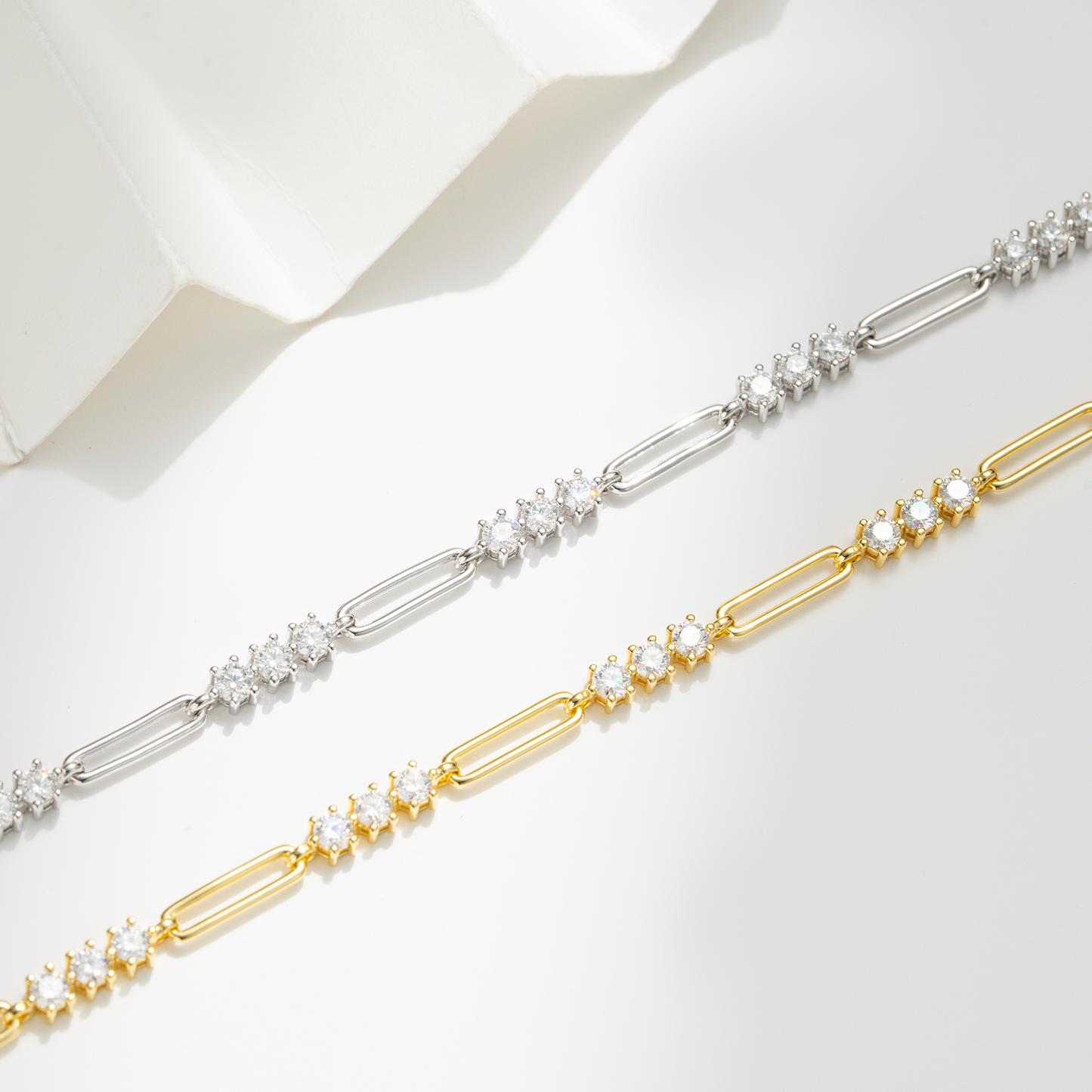 Two paperclip-link chain bracelets, one in silver and one in yellow gold, both adorned with small clusters of clear round-cut stones, displayed diagonally on a clean white surface.