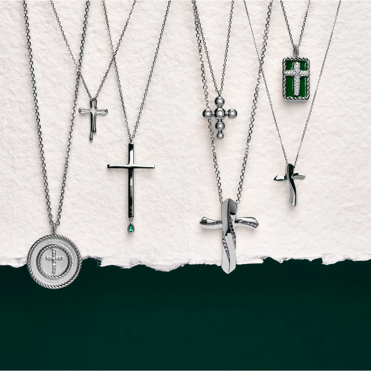A collection of silver-toned cross necklaces with various pendant designs, including a round medallion, a cross with a small green stone, a cross with a small green teardrop, and a cross with a rectangular green stone, arranged against a torn-paper style background with a dark green lower section.