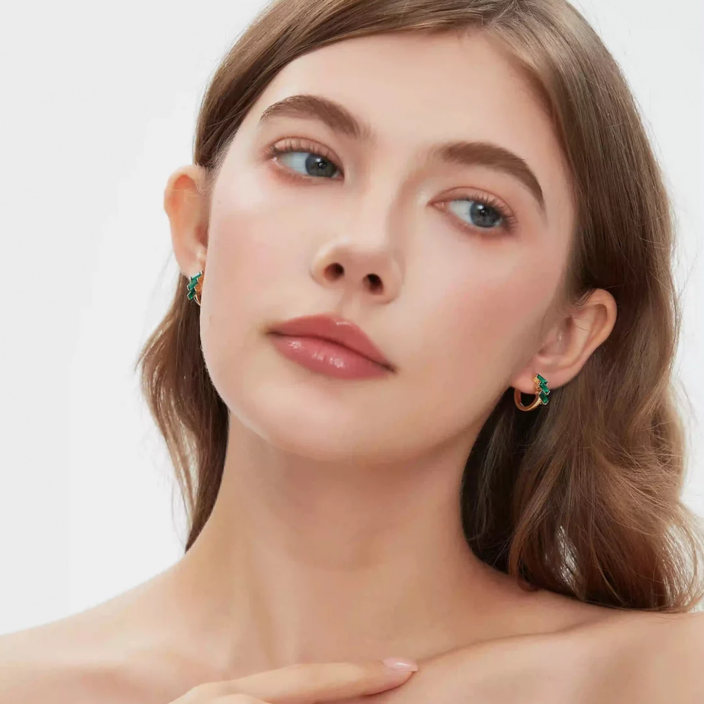 Close-up of a woman wearing small gold crescent-shaped hoop earrings with green stone accents, shown against a clean light gray background.
