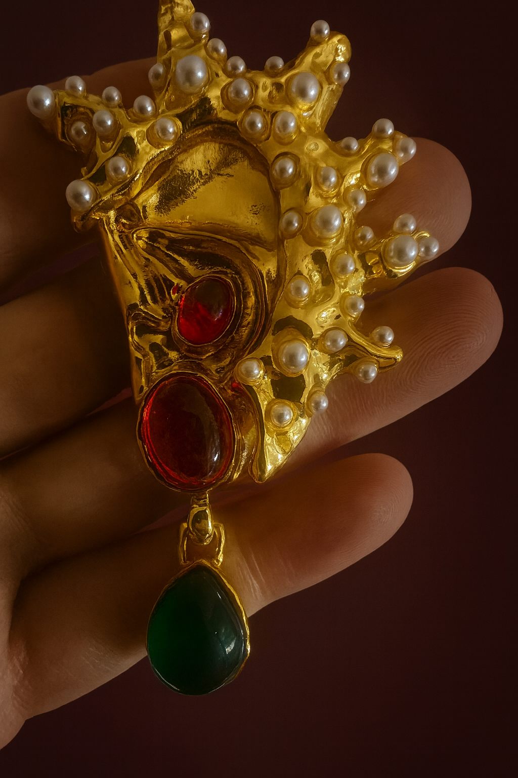 Gold pendant with red and green stones held in a hand against a dark background