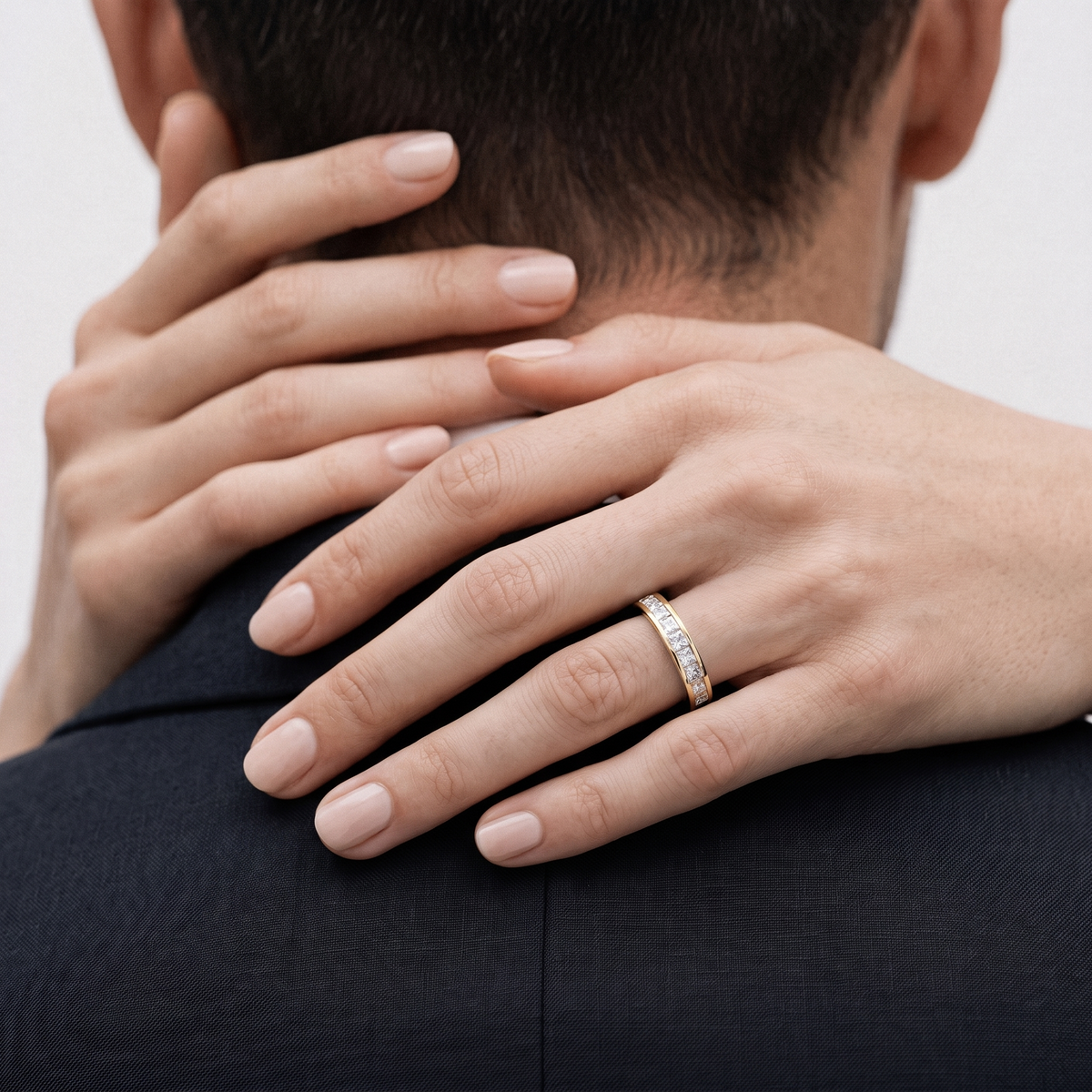 A close-up lifestyle shot of a woman's hand resting on a man's neck, wearing a slim gold band ring with small clear stones on her ring finger.
